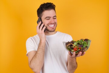 Portrait of successful joyful young handsome Caucasian man holding a salad bowl against yellow wall talking on mobile phone with friend. Lifestyle and communication concept