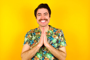 young handsome Caucasian man wearing Hawaiian shirt against yellow wall keeps palms together, has...