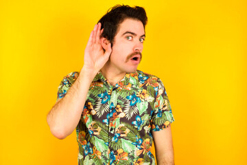 Oh my God! Funny astonished young handsome Caucasian man wearing Hawaiian shirt against yellow wall opening mouth widely looking aside, with hand near ear trying to listen to gossips.