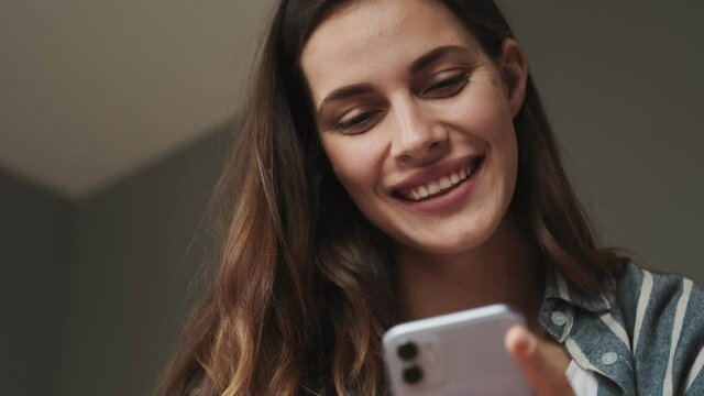 A close-up view of a smiling young woman is using her smartphone sitting in the bedroom at home