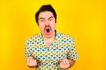 Joyful excited lucky young handsome Caucasian man wearing Hawaiian shirt against yellow wall cheering, celebrating success, screaming yes with clenched fists