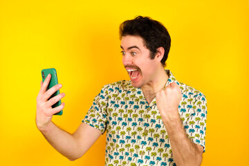 Happy cheerful young handsome Caucasian man wearing Hawaiian shirt against yellow wall receiving good news via e-mail and celebrating success while standing and looking at mobile phone.