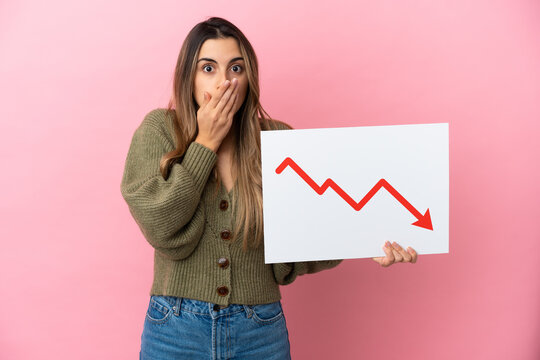 Young caucasian woman isolated on pink background holding a sign with a decreasing statistics arrow symbol with surprised expression