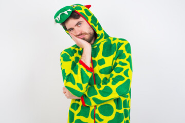 Very bored young caucasian man wearing a pyjama standing against white wall holding hand on cheek while support it with another crossed hand, looking tired and sick,