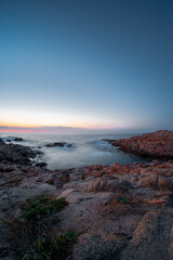 Long exposure photo of sea water washing jagged coastal rocks, Isola Rossa, Sardinia, Italy. Colourful sky after sunset.