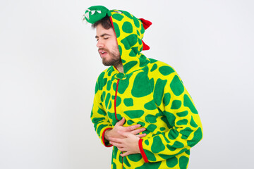 young caucasian man wearing a pajama standing against white wall with hand on stomach because nausea, painful disease feeling unwell. Ache concept.