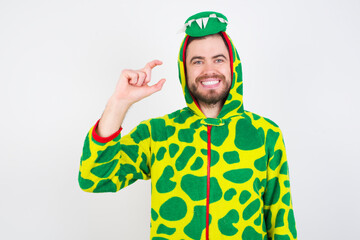 young caucasian man wearing a pajama standing against white wall smiling and confident gesturing with hand doing small size sign with fingers looking and the camera. Measure concept