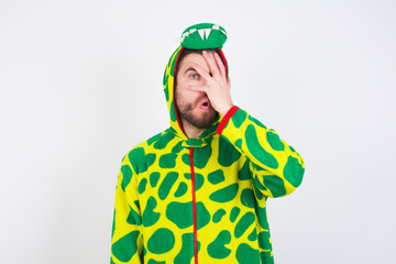 young caucasian man wearing a pajama standing against white background peeking in shock covering face and eyes with hand, looking through fingers with embarrassed expression.