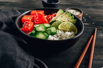 Hawaiian salmon poke bowl with cucumber, avocado and sesame seeds on black wooden table.