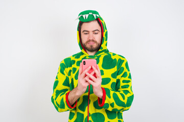 Fototapeta premium Excited young caucasian man wearing a pajama standing against white background winking and eye hold smart phone use read social network news