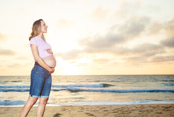 Happy calm pregnant woman during vacations on the sand beach touch belly view over ocean sunset