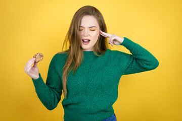 Young beautiful woman eating chocolate cookie over yellow background covering ears with fingers...