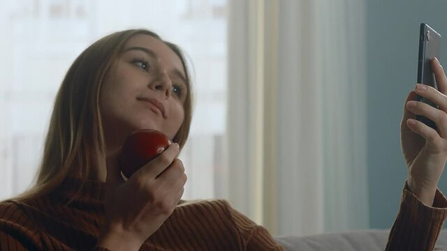 Young girl in blue jeans and brown sweater sits on beige sofa on window background, holding red apple and taking selfie. Close-up of a static plan.
