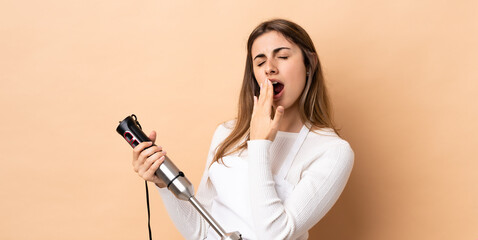 Obraz na płótnie Canvas Woman using hand blender over isolated background yawning and covering wide open mouth with hand