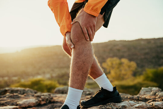 Caucasian Male Athlete Running In Wilderness Holding Hands Around Injured Knee In Pain