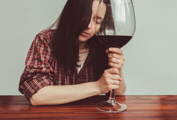 A young girl in a red plaid shirt is tenderly holding a large glass of red wine, closing her eyes. Concept of alcohol abuse, alcoholism, hangover, loneliness and depression. Gray background.