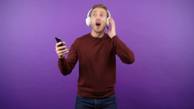 young graceful guy dynamically dancing and sings loudly on isolated purple background in big white headphones