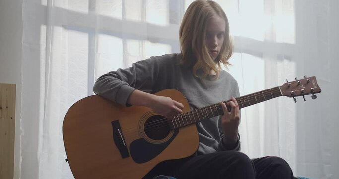 A young girl with blonde hair is enjoying her melody on an acoustic guitar with beautiful sunlight in a white room.