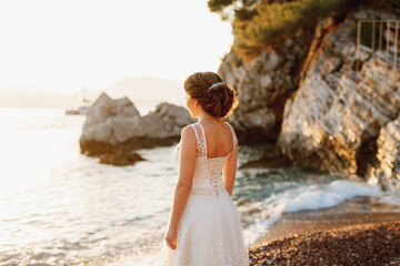 Fototapeta na wymiar A bride in a delicate dress stands on a pebble shore near a cliff and looks into the distance 