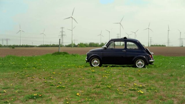 A fiat 500 car parked on a grassy field with windwheels on a cloudy day, tracking shot