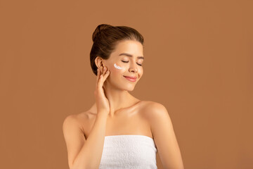 Obraz premium Beauty and wellness concept. Lovely young woman applying face cream over brown studio background