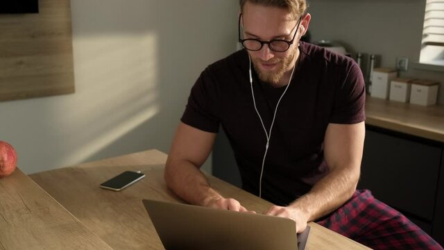 An attractive young blonde man wearing earphones and eyeglasses is using his laptop computer at home