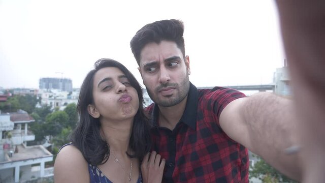 Happy young Indian couple standing on rooftop and posing for funny selfie.Happy girl and cheerful guy having fun making funny faces while taking selfies on smart phone.funny couple making selfie video