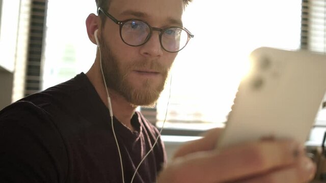 A positive young man wearing glasses and wired earphones is using his smartphone sitting at home