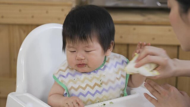 close up irritated and upset asian baby is resisting her mother from wiping the food away from her mouth with a napkin on chair after meal at home.