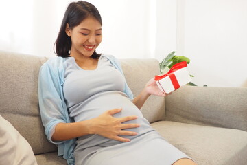 Beautiful Asian mother sitting on sofa at home looking and touching her baby bump with a gift box wrapped with red ribbon in her hand Happy mom smiling and expecting baby with cute present concept