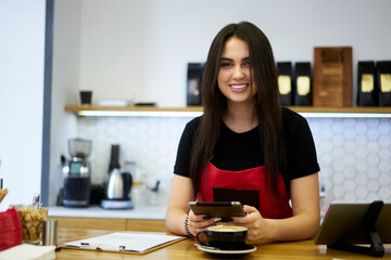 Half length portrait of cheerful prosperous brunette woman barista enjoying work in coffee shop making orders via app on digital tablet, smiling female waitress at bar holding cashier touchpad