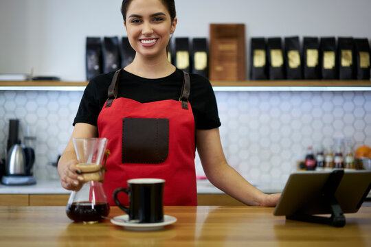 Half length portrait of cheerful caucasian woman 20s bariste in uniform standing at bar in cafe using glass filter for making aroma coffee beverage, smiling prosperous female bariste using jar