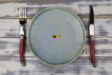 Top view on dish with one isolated red yellow capsule, cutlery on wood table - medication intake...