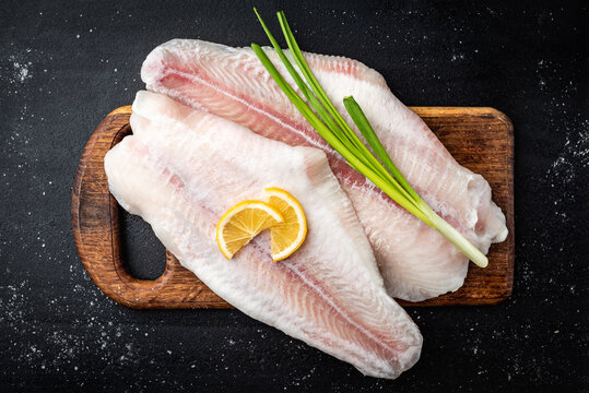 Fillet of raw pangasius fish with lemon and green onion on cutting board on dark background. Top view.