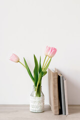 A bouquet of three pink tulips stands in a glass vase with lace. Next to it are notebooks or notepads and a mug of tea, a photo album in a velour fabric cover of beige color