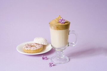 Beautiful dalgona drink a frothy coffee in a transparent mug and flowers of lilac on a purple background. Sweets on a plate: the donut and sweet marshmallows