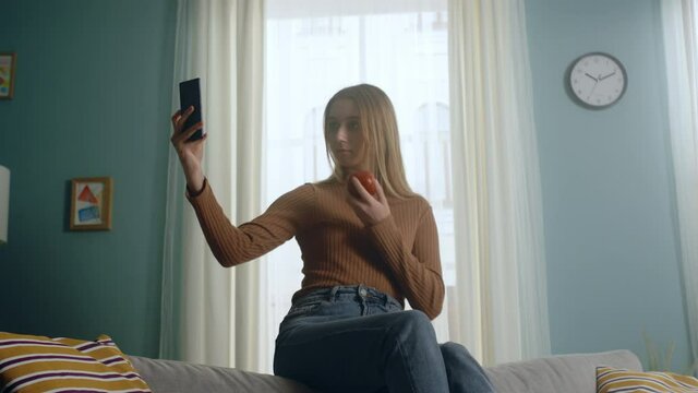 Young woman in blue jeans and brown sweater sits on back of sofa on background of window, holding red apple and taking selfie. Clock hangs on wall. Average plan.