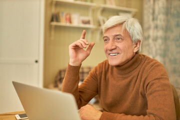 Portrait of handsome positive mature male in warm sweater working from home using portable computer for distant work, having bright idea to promote his business online, raising index finger