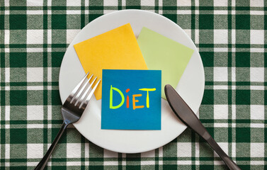 A fork and knife, white plate with blue sheet of paper which the word Diet is written stand on a checkered green tablecloth.The concept of a balanced diet, ration and medical fasting. Copy space.