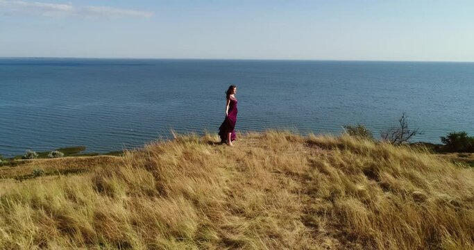 Young woman stands by the sea in burgundy dress in summer. Brunette walks on dry grass near the ocean