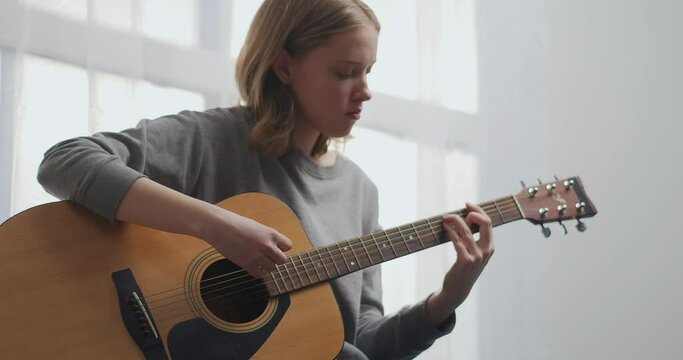A teenager plays guitar in a white living room with beautiful sunlight. Woman composes music indoors. A girl is practicing a musical instrument alone during a pandemic.