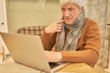 Unhealthy frustrated mature man with gray hair sitting at desk wrapped in scarf having sore throat, feeling sick, trying to consecrate on working task, using wireless internet connection on laptop