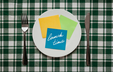A fork and knife, white plate with blue sheet of paper which the word Lunch time is written stand on a checkered green tablecloth.The concept of a balanced diet, ration and medical fasting. Copy space