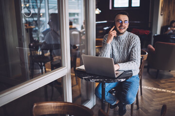 Smiling young male freelancer in glasses talking on smartphone while using laptop in cafe