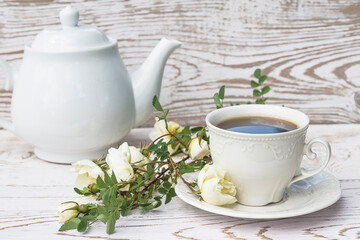 Coffee break in retro style. White cup of coffee or tea, white teapot and branch with white wild roses flowers on white paint rustic wooden background