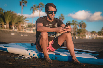 a young man uses his mobile phone sitting on his paddle board on the beach