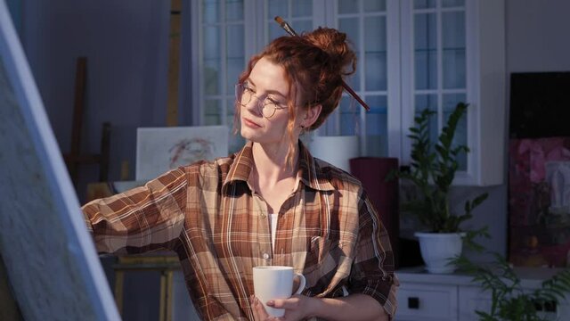 art female with paintbrush in hair enjoying a hot coffee after the creative process, happy young woman with a cup in hands looking at a large canvas in studio