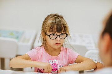 Naklejka na ściany i meble Young cute girl with down syndrome in pink shirt and black glasses sitting at white desk and looking to her teacher.