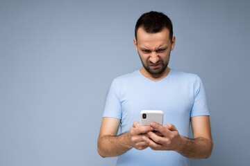 Photo of dissatisfied handsome young man with beard wearing everyday blue t-shirt isolated over blue background holding and using mobile phone communication online on the internet looking at gadjet