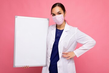 Young beautiful woman nurse in protective face mask holding an empty magnetic board isolated on pink background